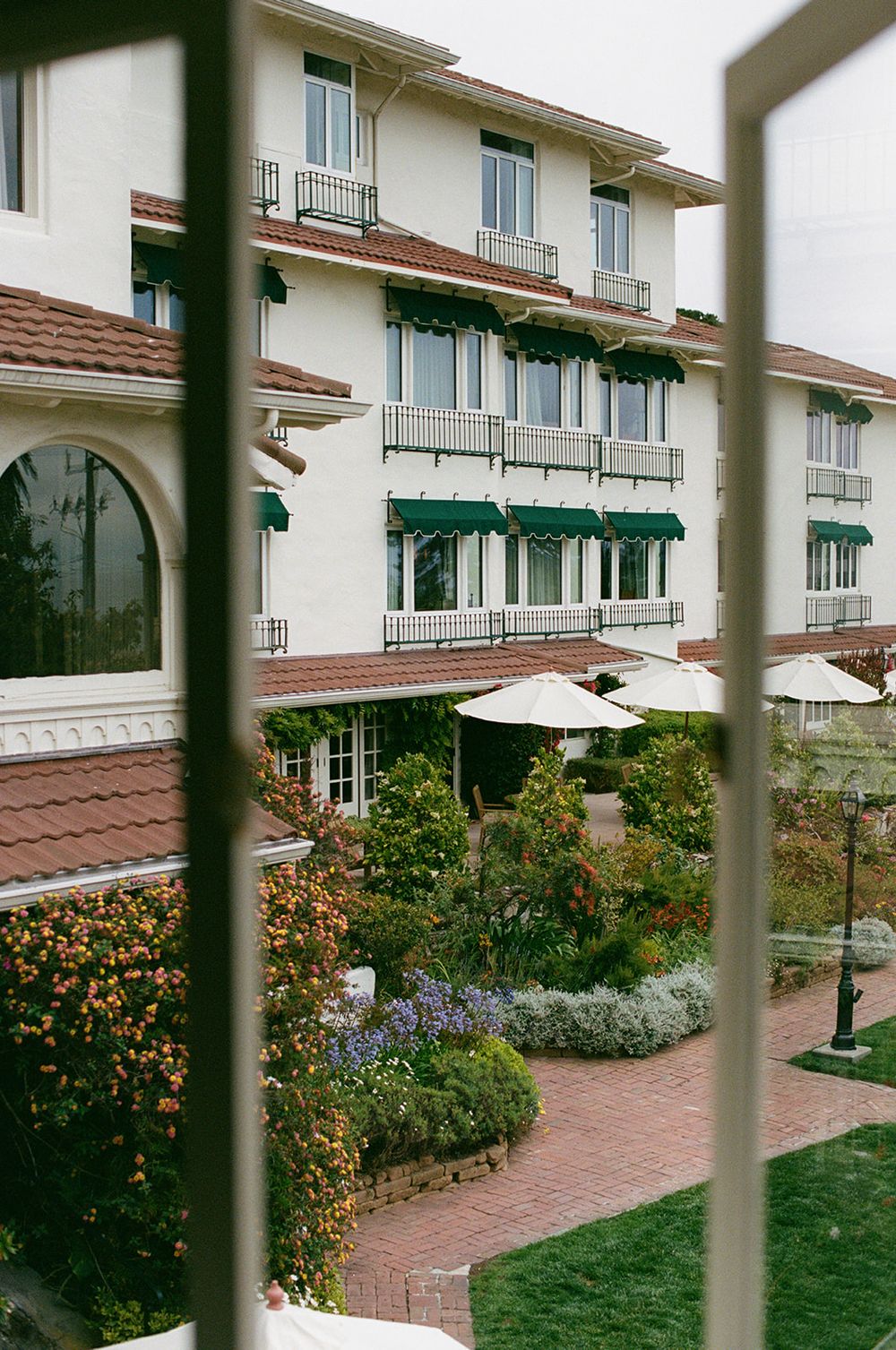 Mediterranean-style building exterior with white walls, red tile roof, and lush garden landscaping along a brick pathway.