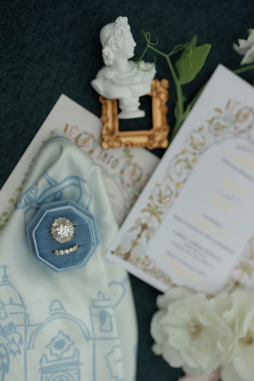 Wedding details flatlay with blue and gold invitations, rings, and white floral accents.