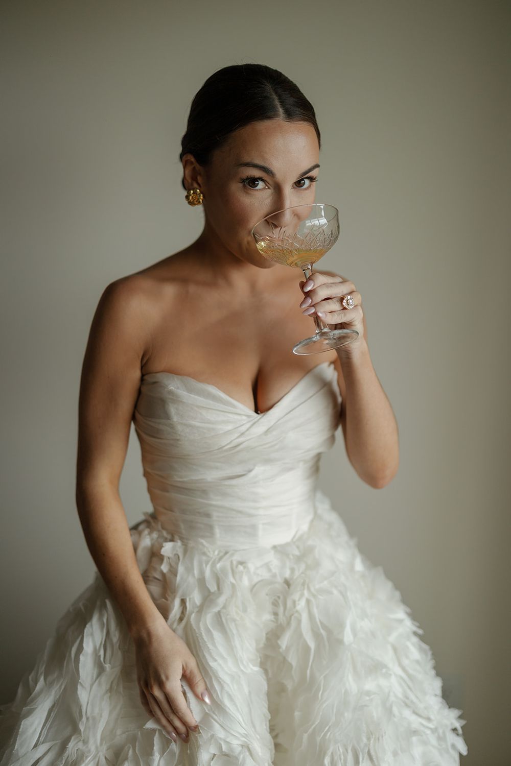 Bride in white ruffled wedding gown holding champagne glass against neutral background.