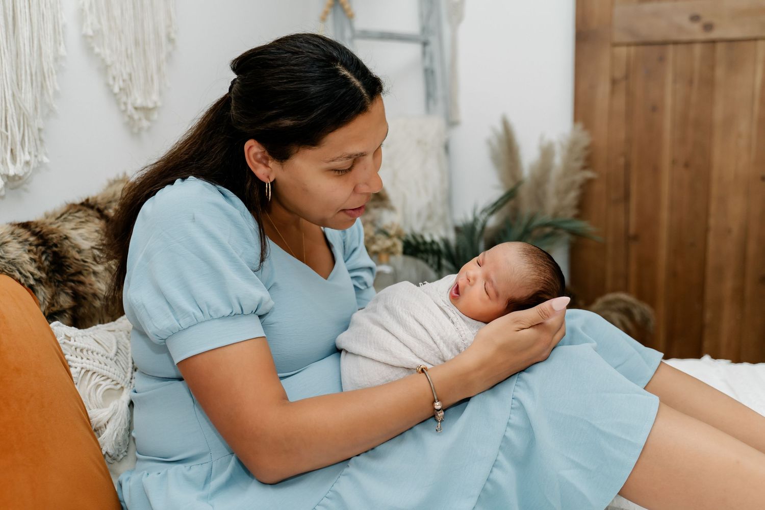 A gentle moment between parent and newborn baby on a couch with cozy blankets and macrame decor in the background.