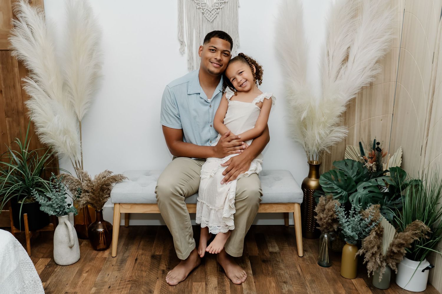 A sweet family photo of a loving pair against a white wall with pampas grass and tropical plants.