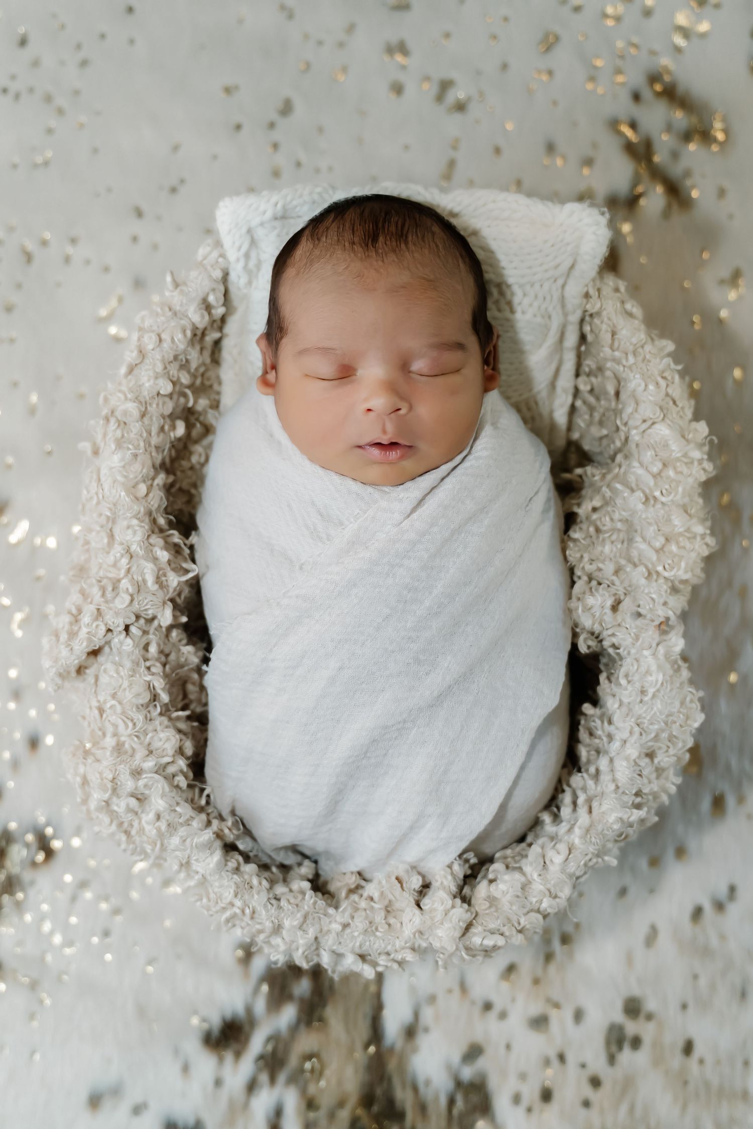 Sleeping newborn swaddled in white blanket nestled in cozy textured fur setting with scattered sparkly accents.