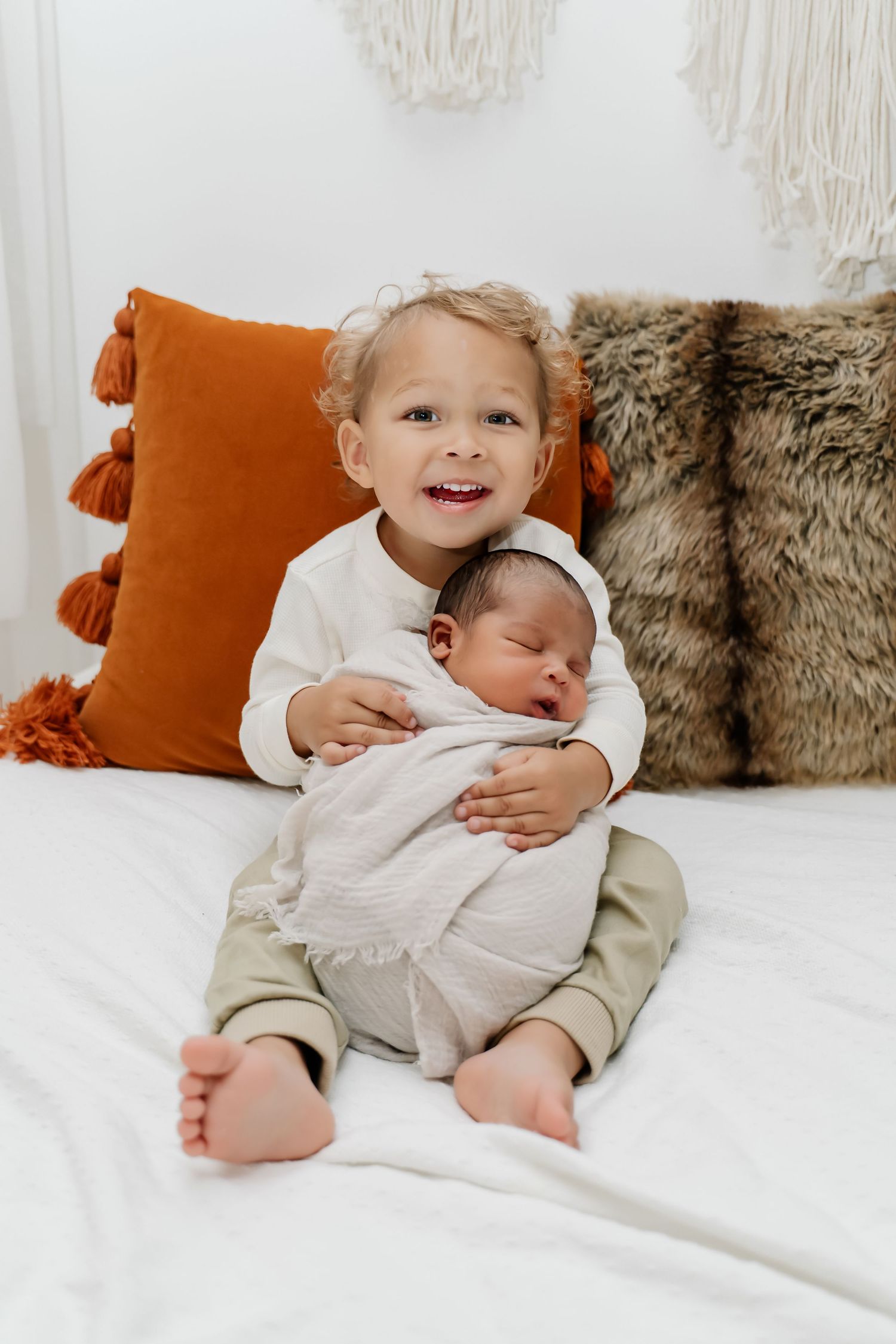 Sweet sibling bonding moment on a cozy white bed with orange pillows and fur throw blankets.
