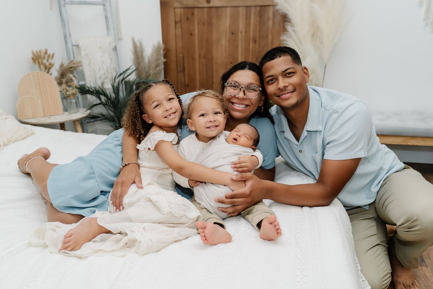 A joyful family lounges together on a white bed, all wearing light-colored clothing against a minimalist backdrop.