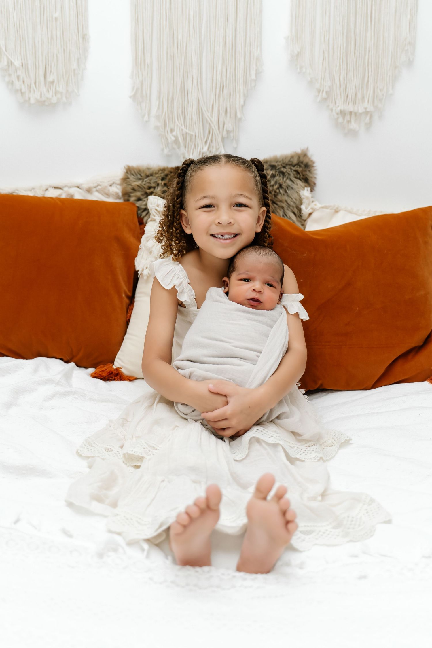 Siblings sitting together on white bedding against orange velvet pillows with macrame wall hanging in background.