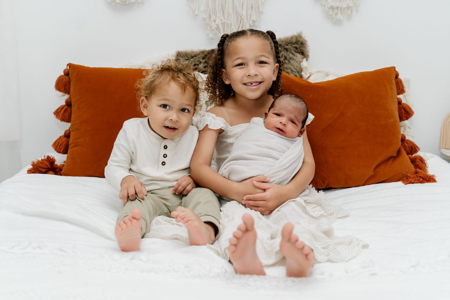 Three young children sitting together on a white bed with orange pillows, smiling and cuddling.
