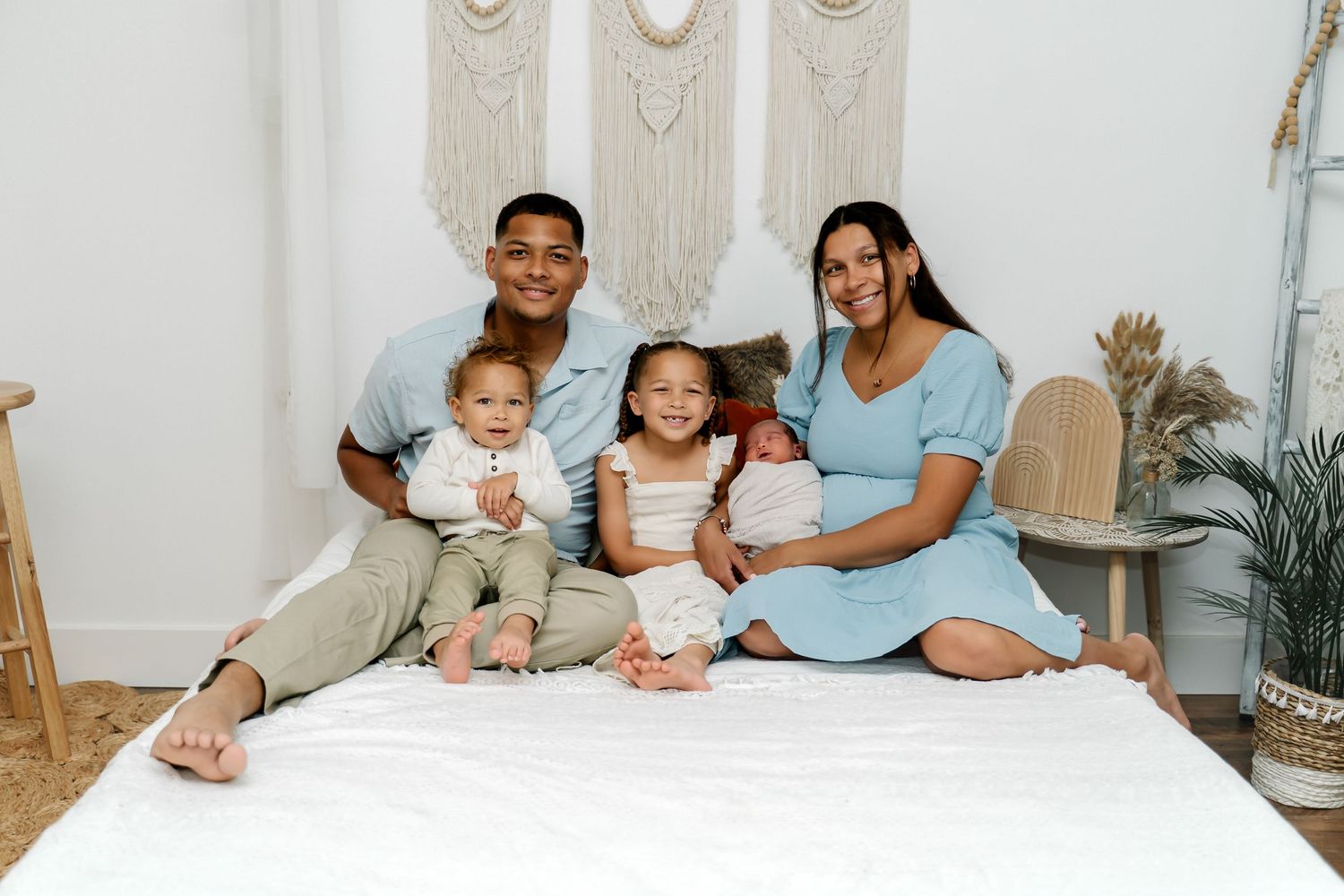 A family of five sits together on white bedding in a cozy bedroom with boho wall decor and plants.