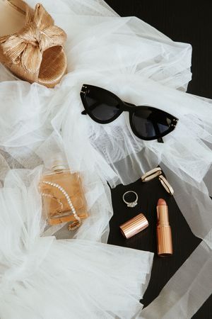 Flatlay of wedding accessories including sunglasses, lipstick, tulle fabric and rose gold bow against dark background.