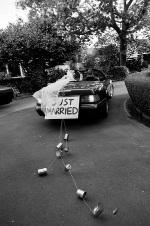 Black and white photo of a car decorated with a "Just Married" sign and trailing tin cans on a driveway.