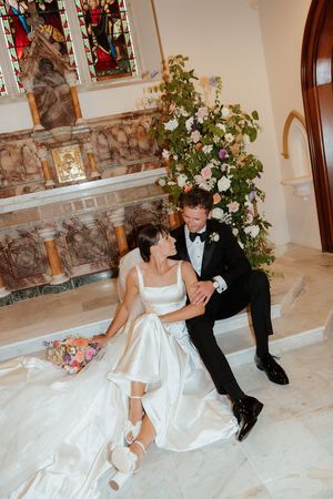 Newlywed couple sits playfully on marble church steps surrounded by elegant floral decorations.