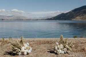 Two white floral arrangements sit on a beach shoreline with calm lake waters and mountains in the background.