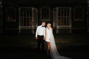 Couple in formal wedding attire poses together at night against a dark gothic architectural backdrop.