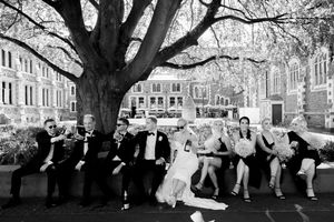 Wedding party sits casually on stone ledge under large tree branches in black and white photograph.