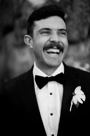 Four black and white photos of someone in a tuxedo with boutonniere smiling joyfully during what appears to be a wedding.