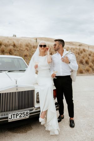 A wedding couple poses playfully near a vintage Rolls Royce on a dusty desert road.