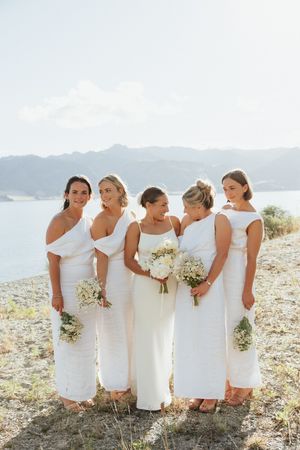 A bridal party wearing elegant white dresses poses together on a cliffside overlooking a scenic mountain lake view.