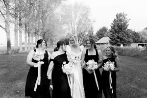 Black and white photo of a wedding party in formal attire standing together on a lawn with trees in the background.