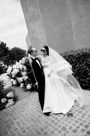 Black and white wedding photograph showing newlyweds embracing outside a building with brick pavement and hydrangeas.