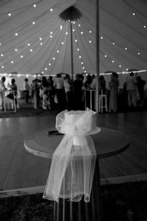 A white tulle bow decorates a folding chair at a wedding reception under a tent with string lights and guests mingling.