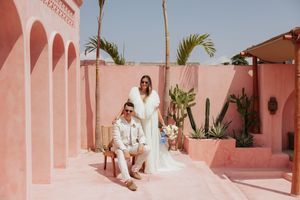 Couple in white attire pose at pink-walled tropical venue with palm trees.