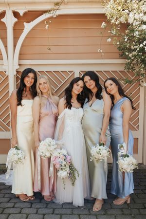 A wedding party in pastel silk dresses gathered in front of a peach-colored wall with white floral decorations.