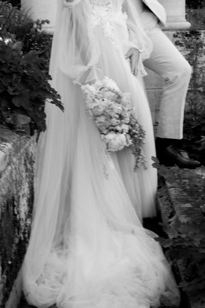 A black and white wedding photograph showing a flowing white bridal gown and bouquet against a garden backdrop.