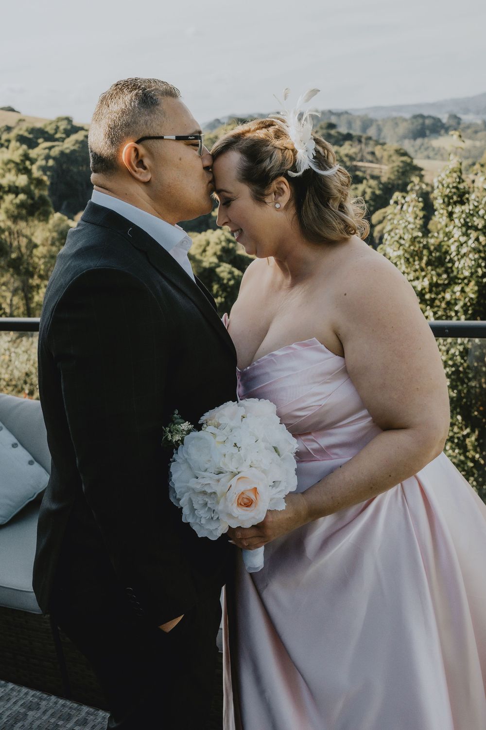 Wedding couple shares an intimate moment on a balcony overlooking scenic hills with bride holding white bouquet.