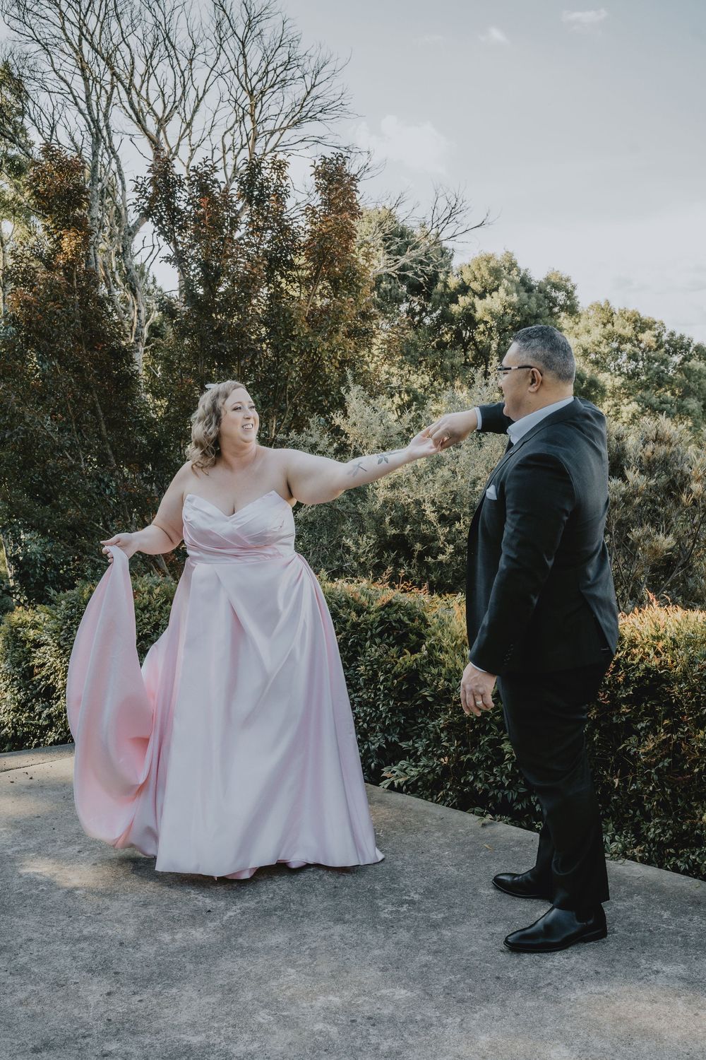 A couple shares a playful moment outdoors, with the person in pink dress gesturing while wearing formal attire.