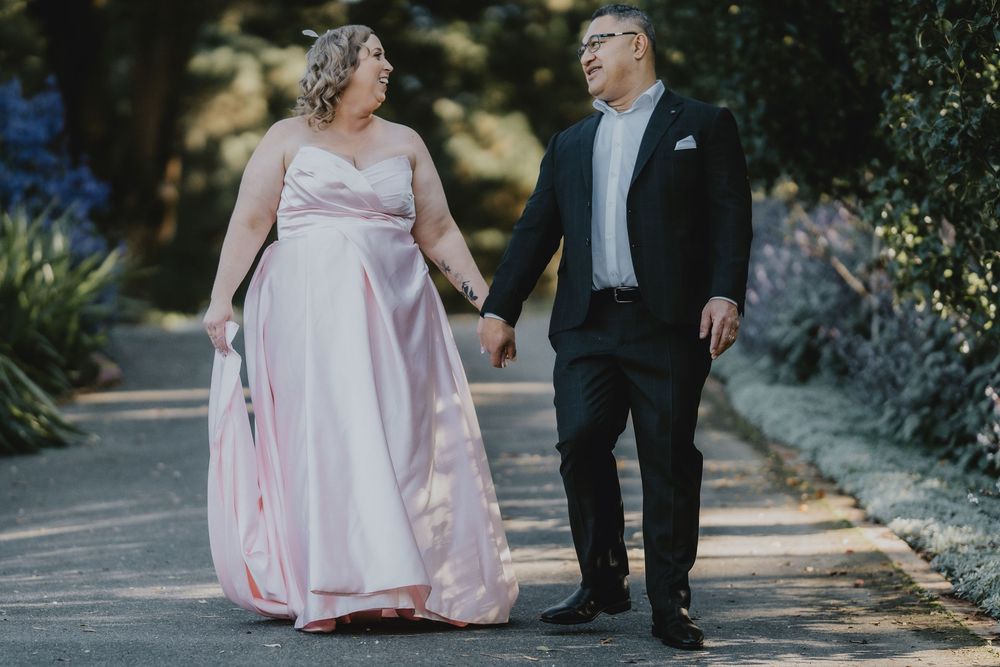 Couple in formal attire walking together on a garden path in soft natural lighting.