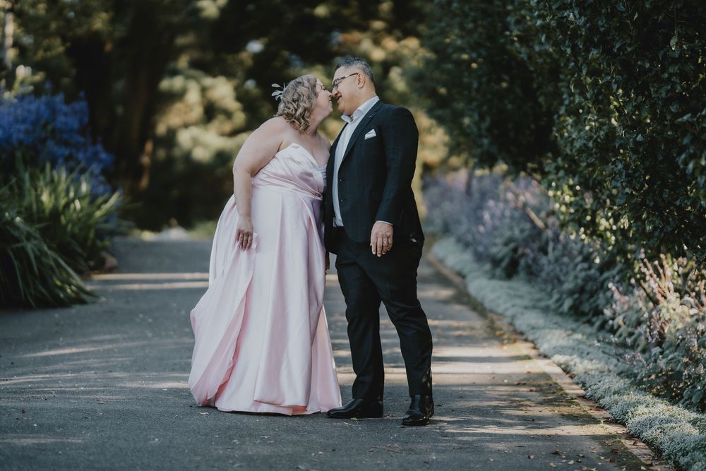 A couple shares a romantic moment on a garden path, with the bride wearing a pink gown and groom in black tuxedo.
