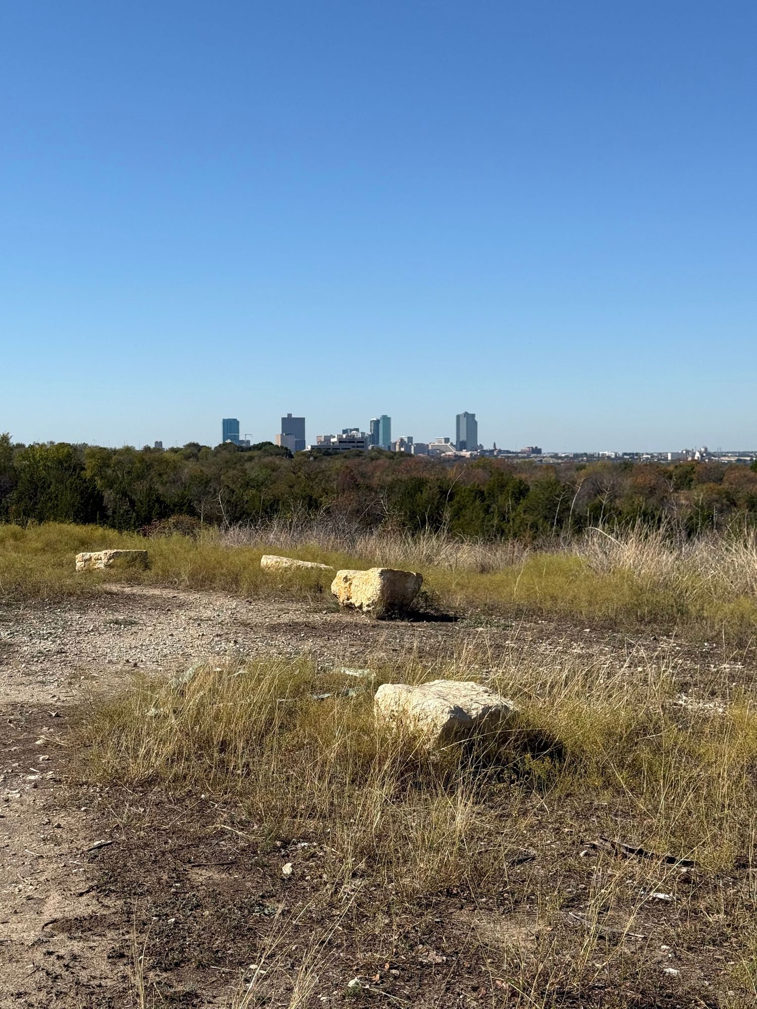 Fort Worth skyline viewed from a hilltop trail with large limestone rocks in the dry grass foreground. Tandy Hills Nature Preserve