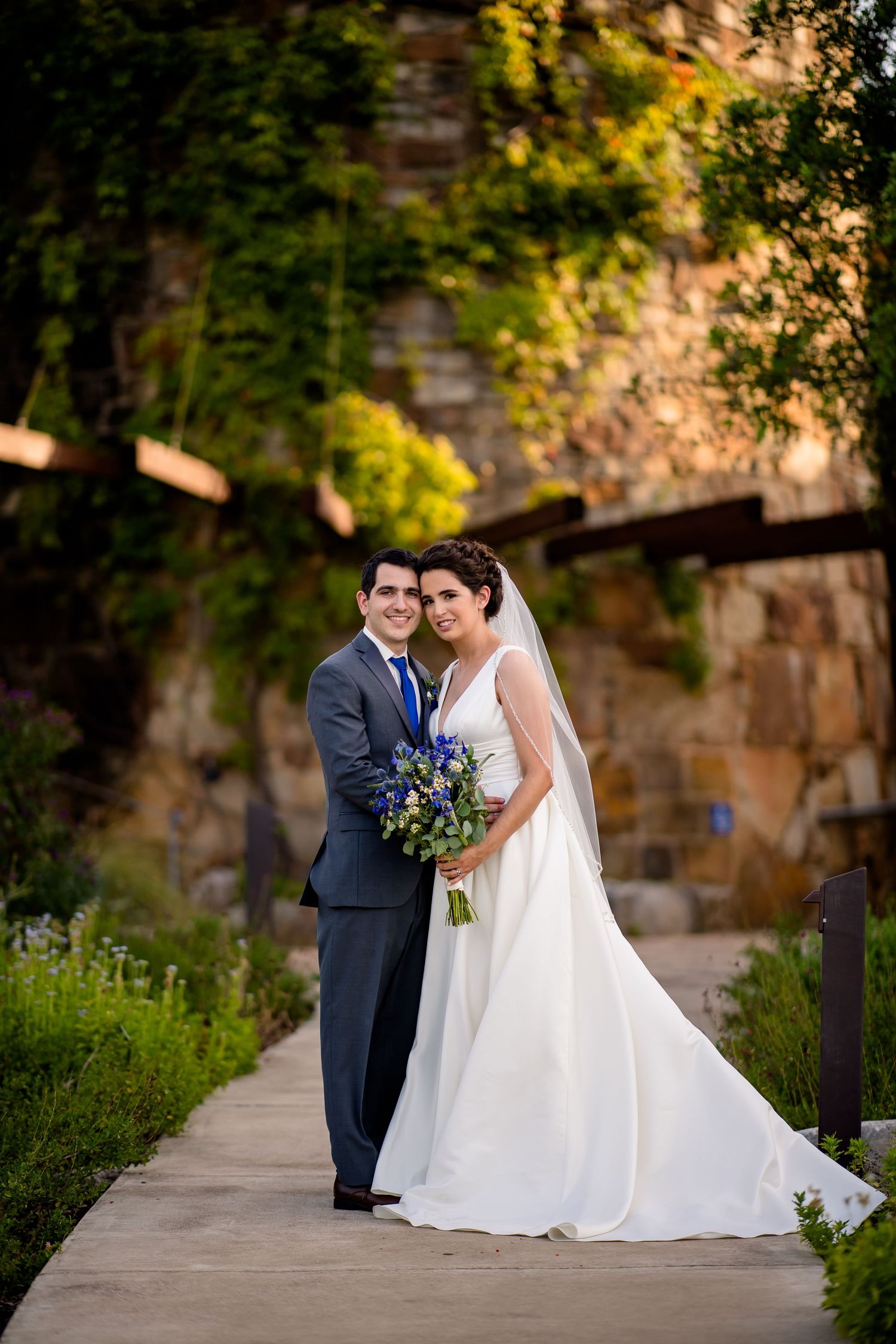 Wedding couple stands together on stone walkway holding blue bouquet with natural rock wall and greenery background.
