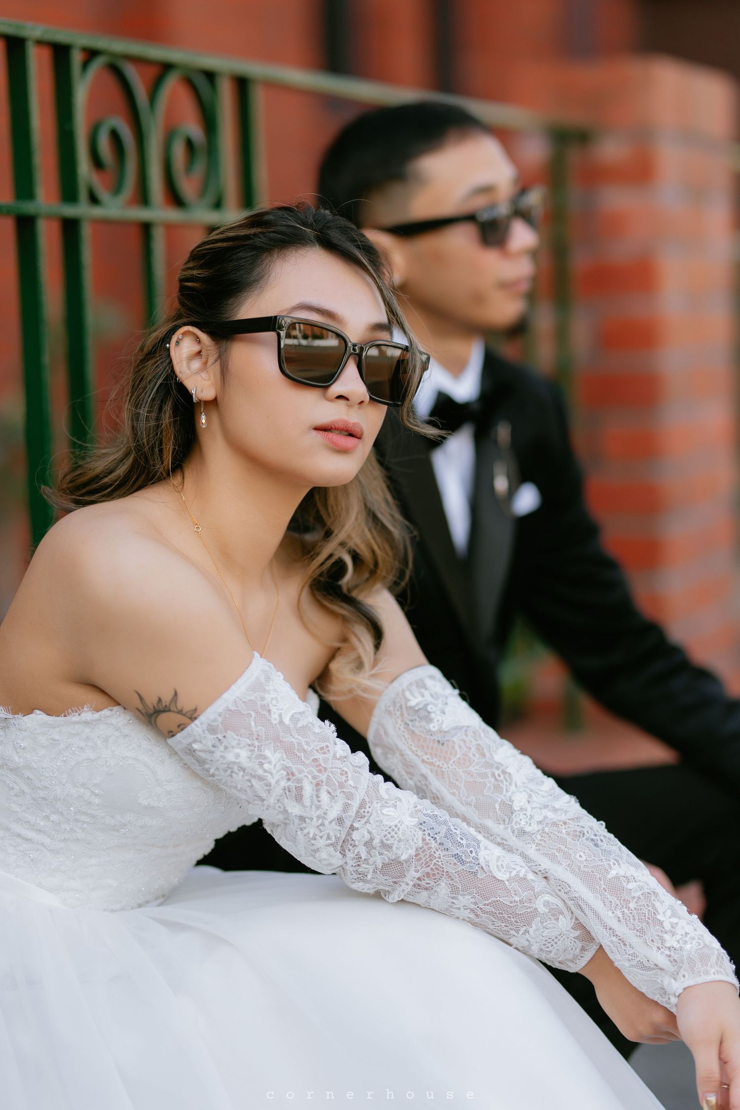 Stylish wedding couple in sunglasses pose against brick wall with ornate green metal railing.
