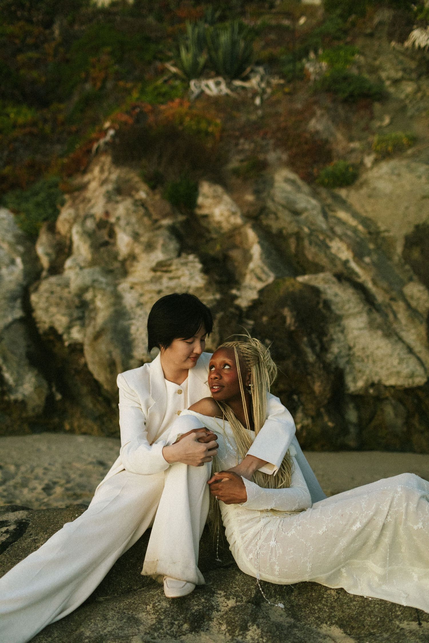 Two brides lean on each other, sitting down on a rock at Laguna Beach, California