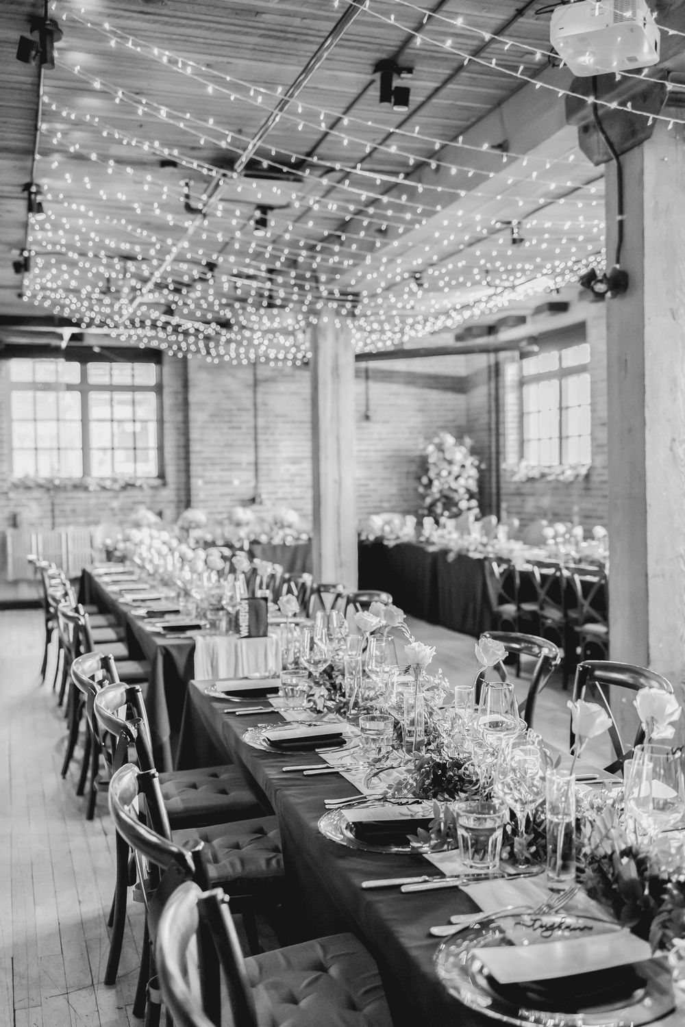 Long reception tables set for an elegant wedding under twinkling string lights in an industrial-style venue with exposed walls.