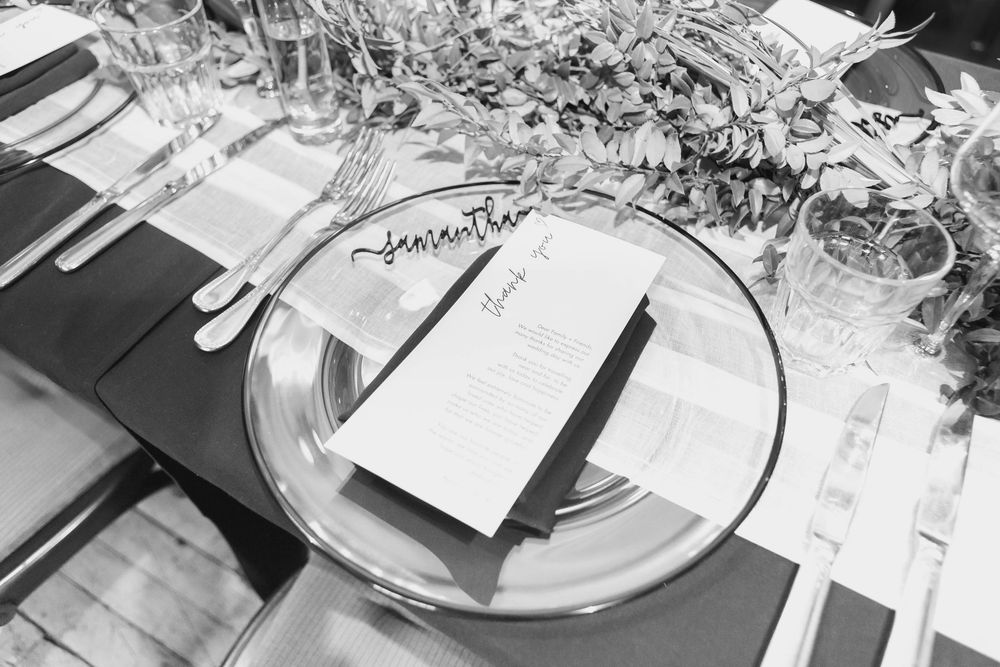 Black and white close-up photo of elegant place settings with wine glasses and formal dinnerware on a white tablecloth.