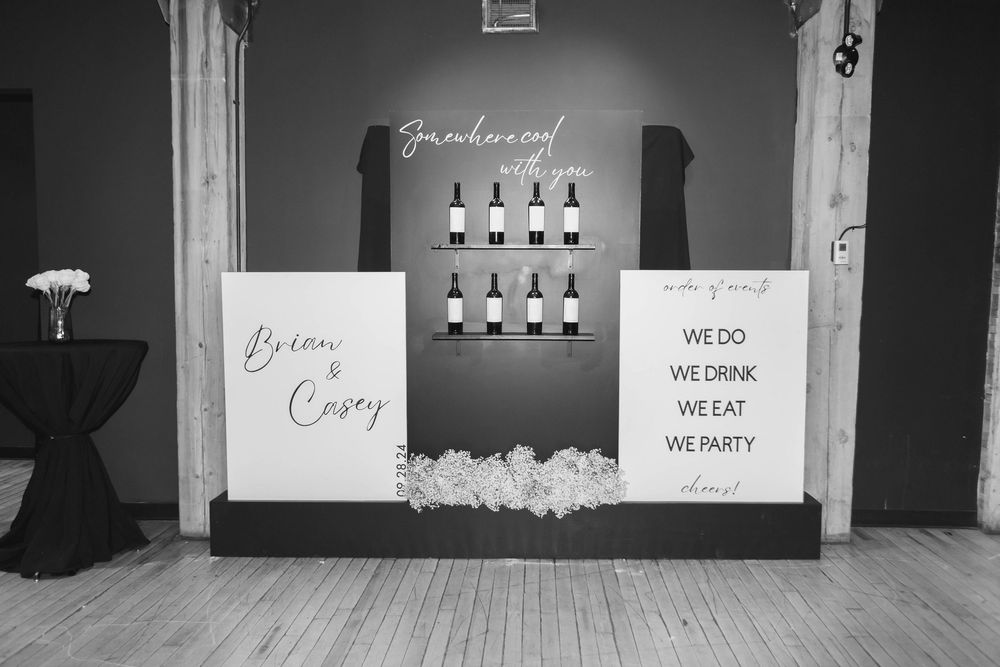A black and white wedding reception display with drink menu board and decorative signs against a dark wall.