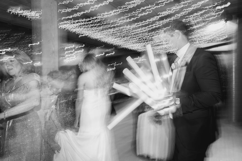 Motion-blurred black and white photograph of people dancing at a celebration with light streaks.