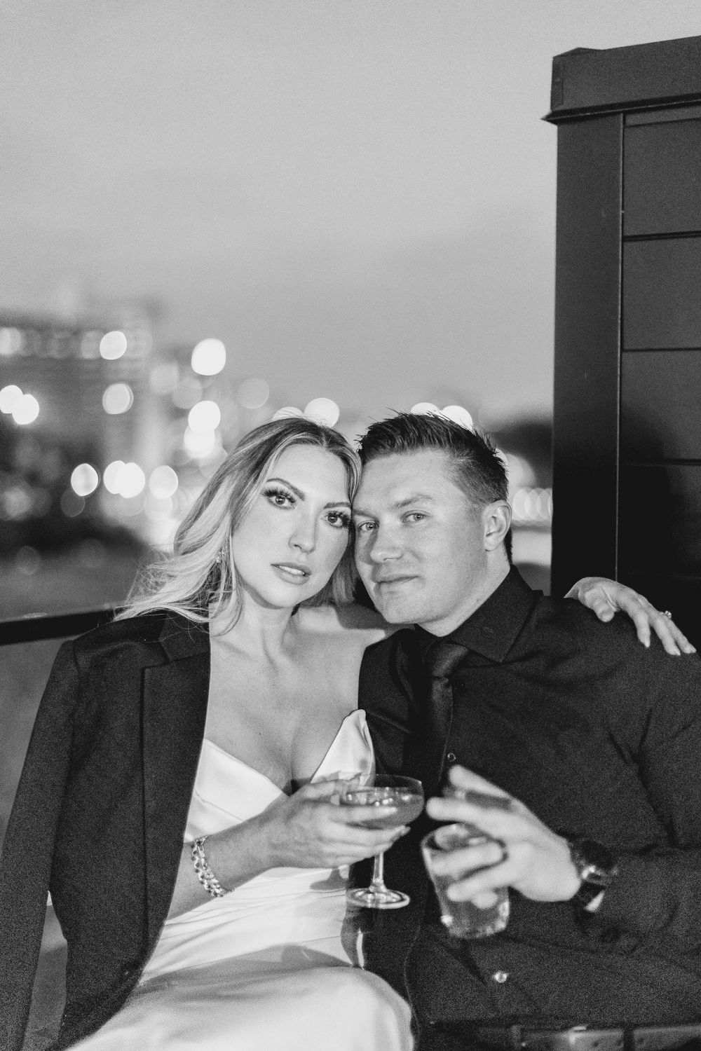 A black and white shot of a couple enjoying drinks on a rooftop with city lights bokeh in the background.