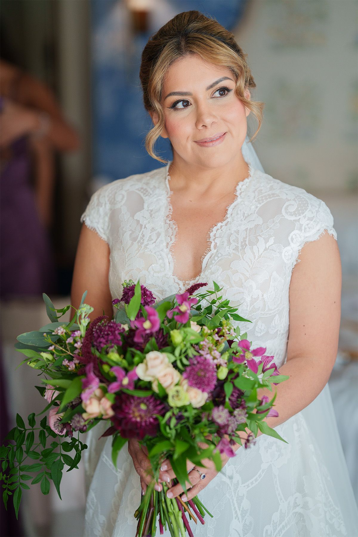 A bride in a delicate white lace gown holds a stunning bouquet of purple and pink flowers with greenery.