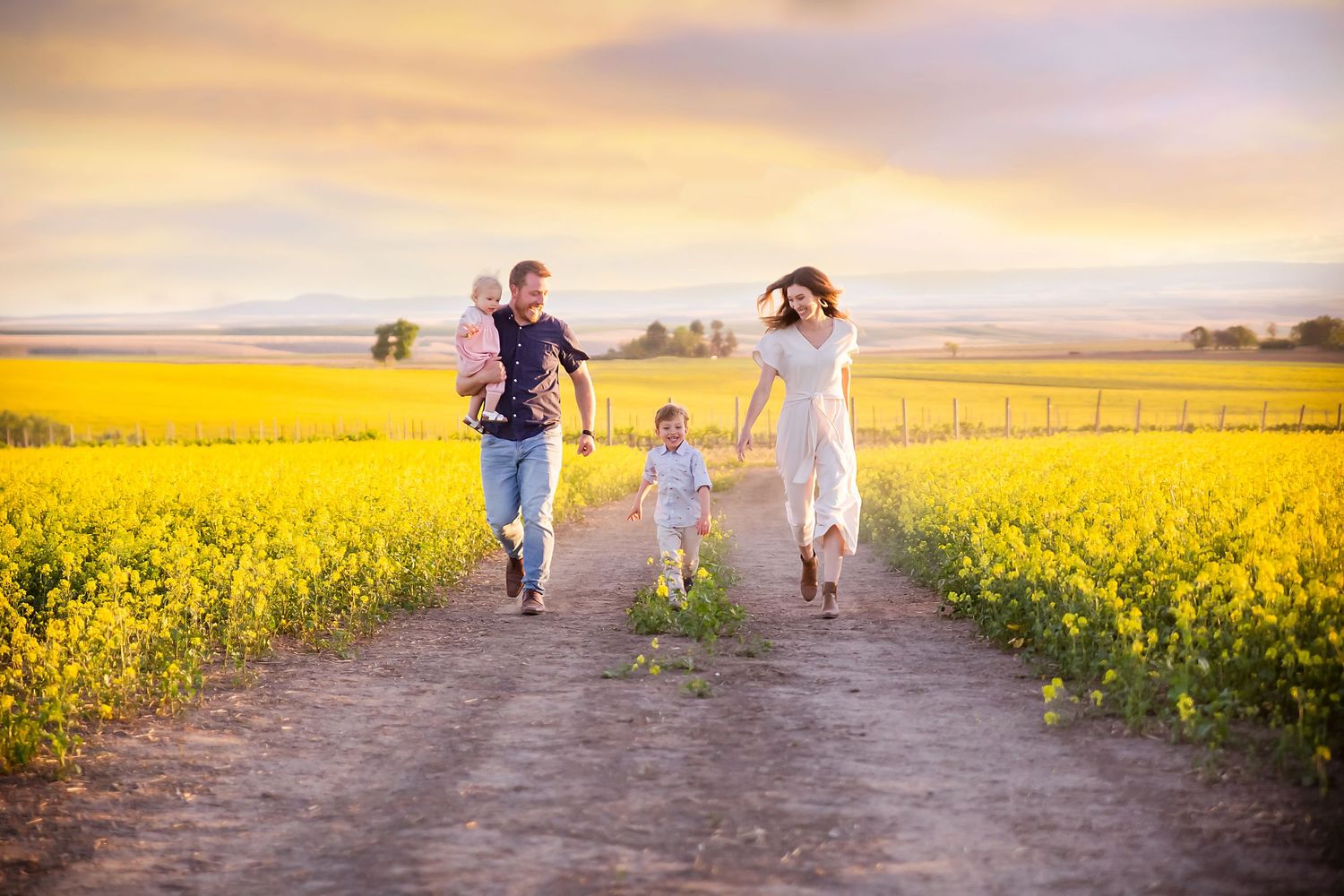 Family running together on dirt path through vibrant yellow canola field at sunset.