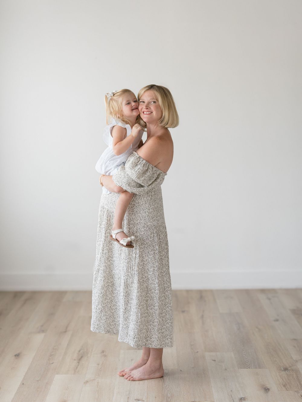 A full body shot of a mother holding her daughter on light wood floors and a airy white background.