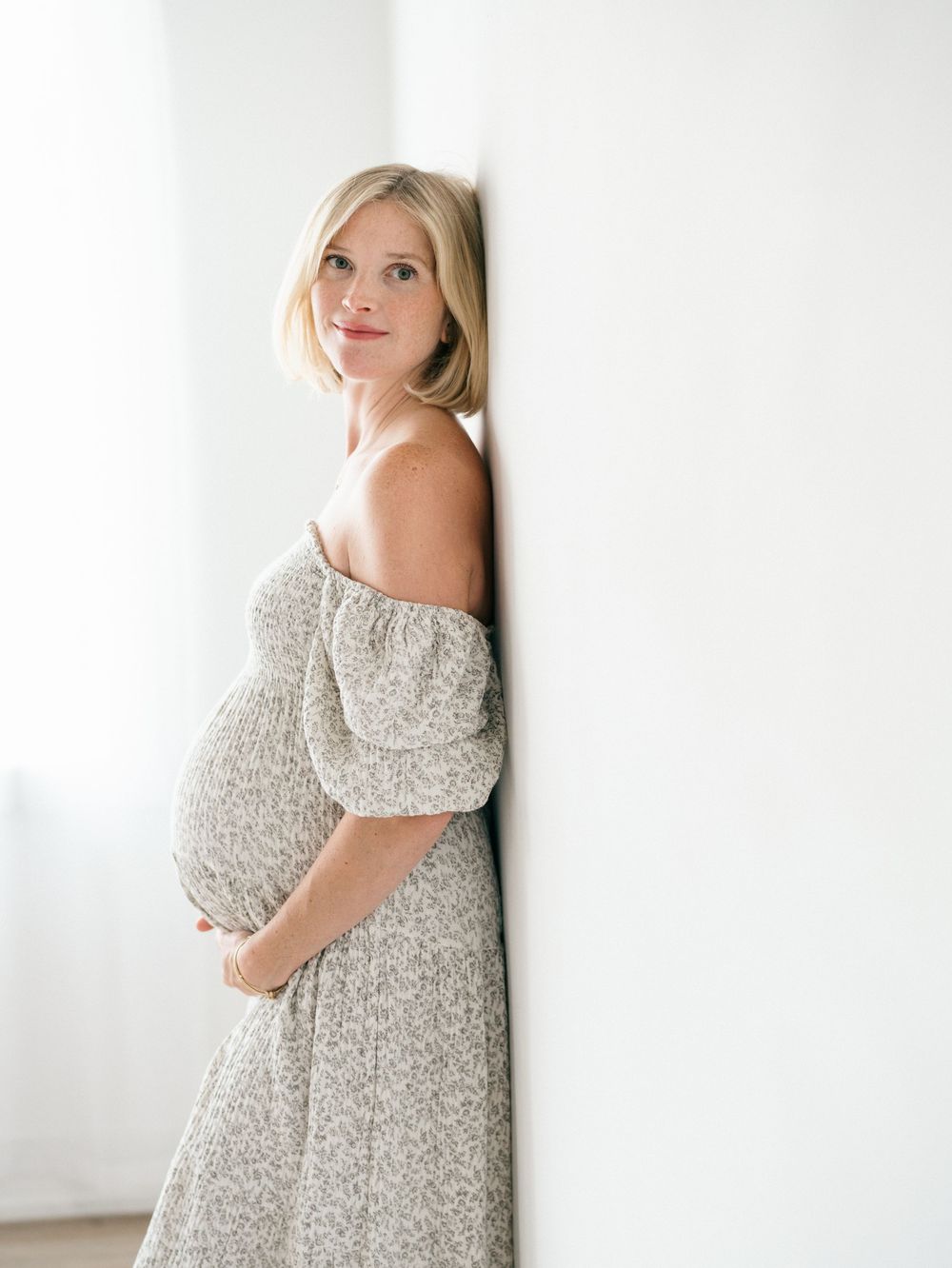 Expecting mother backlit while standing barefoot in a light-filled studio, wrapped in a flowing white and gray floral nothing fits but dress