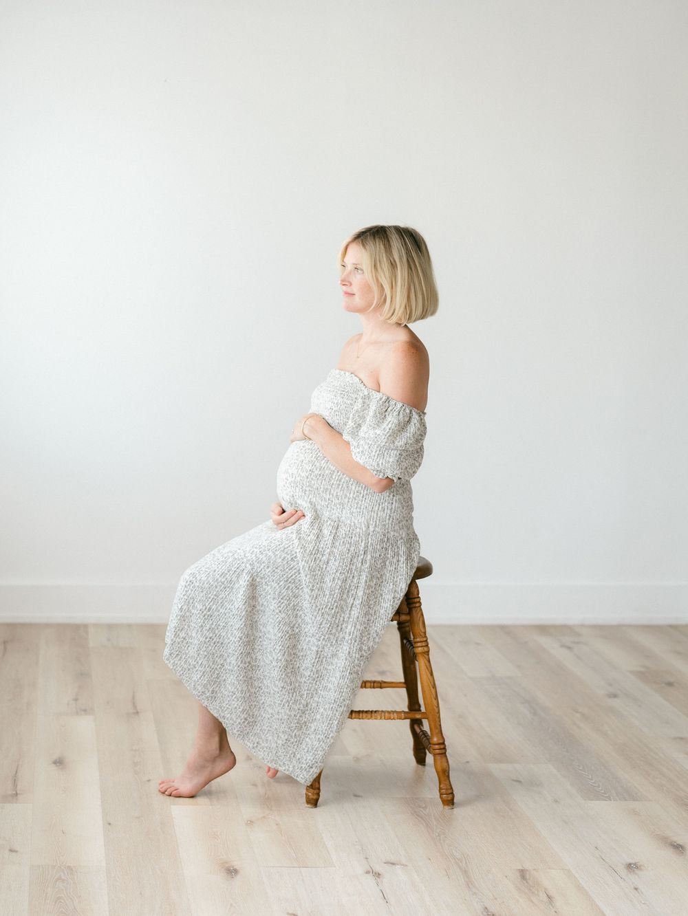 A pregnant woman sits on an antique wood stool looking out the window.