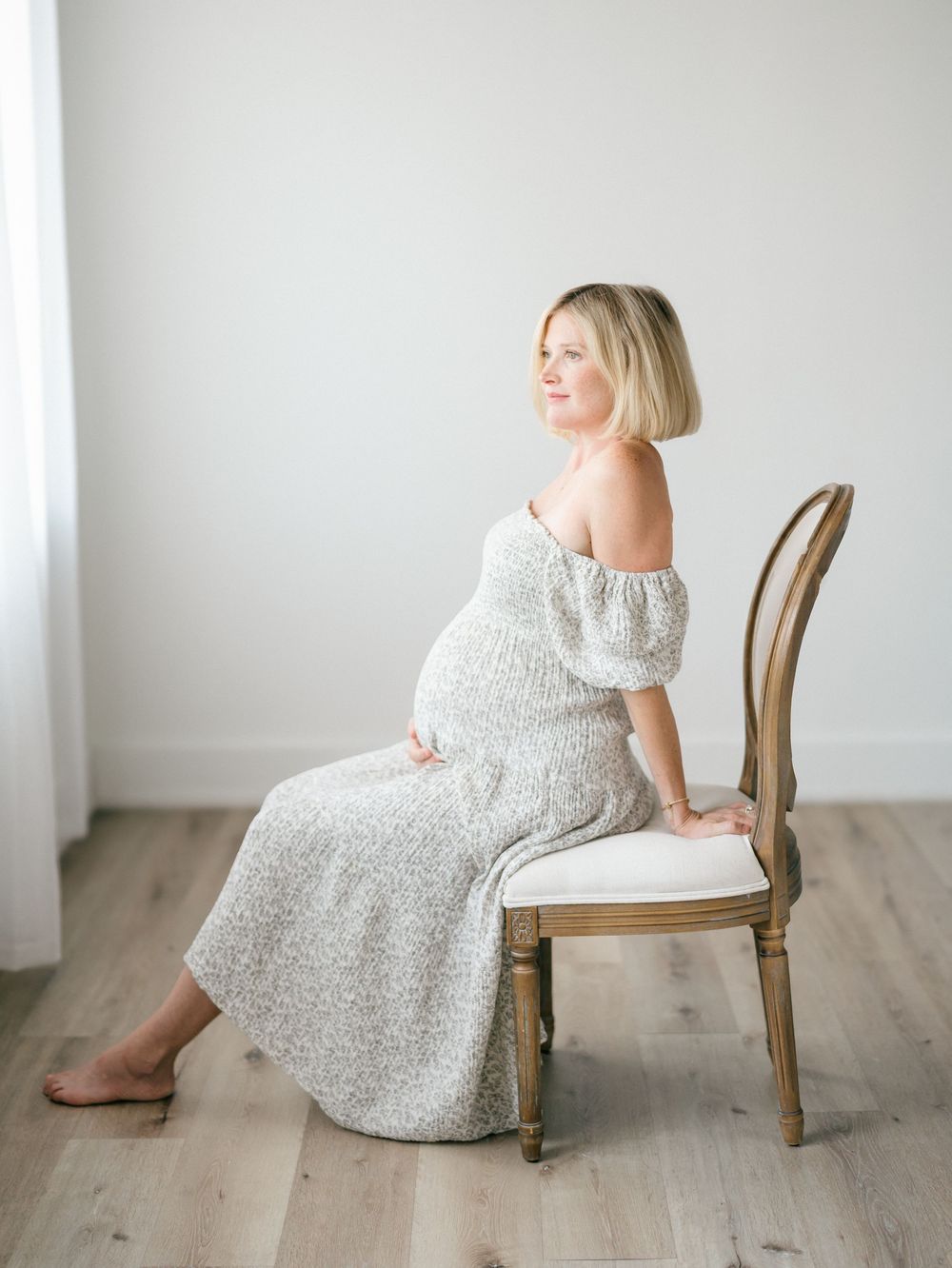 Pregnant woman in a flowing floral dress cradling her belly on a chair during a studio maternity session in Monmouth County, NJ.