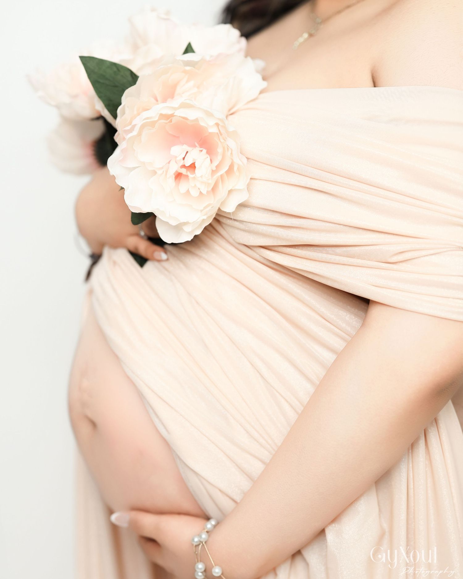 A pregnant figure in a blush pink pleated gown holds delicate white and peach flowers against a light background.
