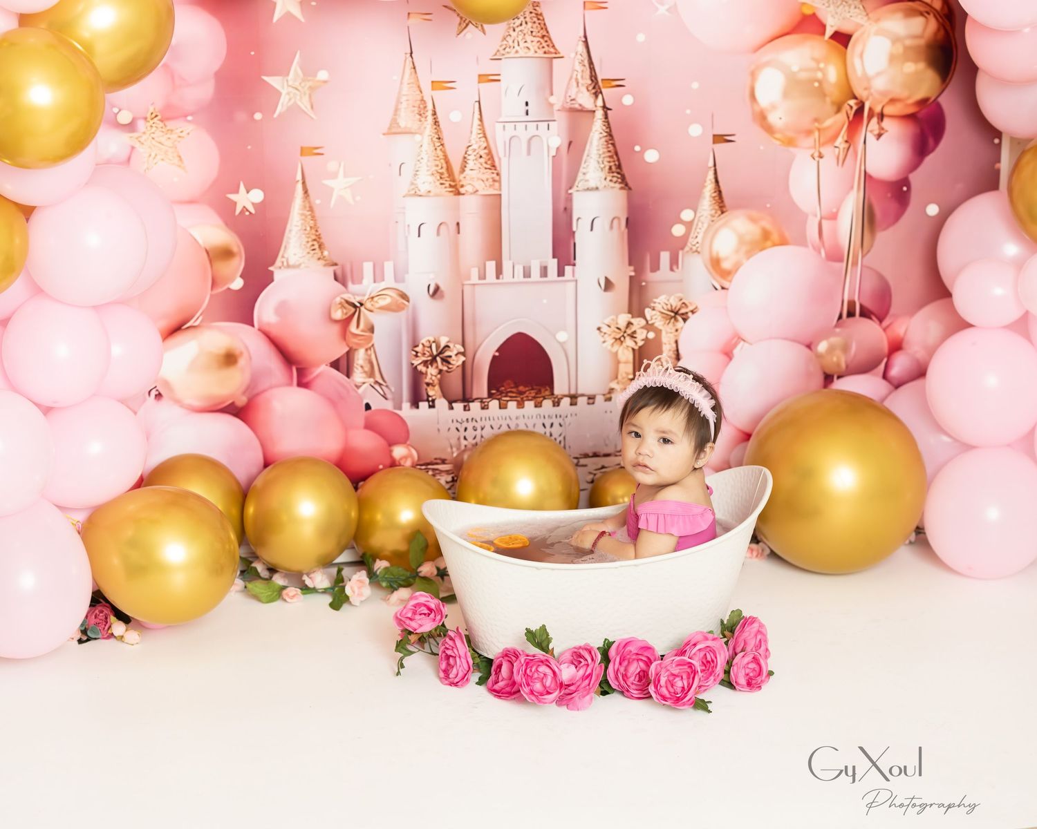 A dreamy cake smash setup with pink and gold balloons surrounding a white bathtub and fairy tale castle backdrop.