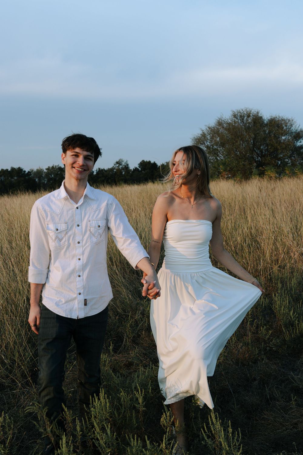 A couple in wedding attire playfully walks through a golden grass field at sunset.