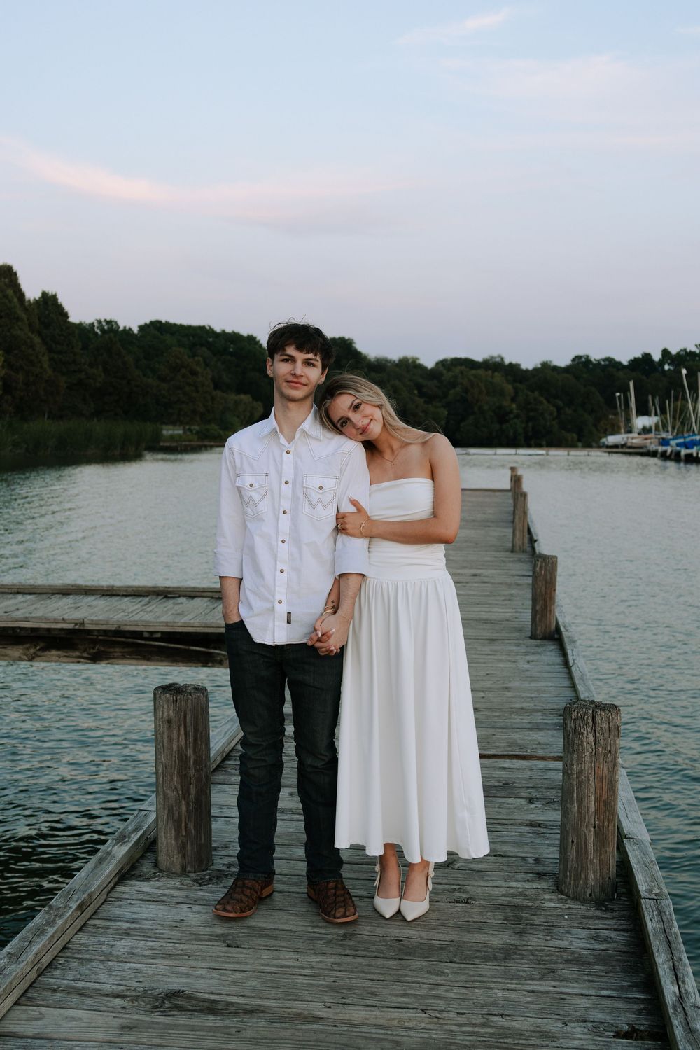 A couple in white and black outfits poses together on a lakeside wooden pier at dusk.