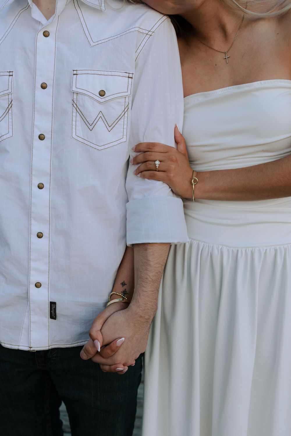 Close-up of couple holding hands, with white western shirt and flowing white strapless dress details visible.
